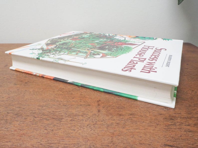 May include: A hardcover book titled "Success With House Plants" by Reader's Digest. The book's cover features a colorful illustration of a greenhouse with various plants. The book is resting on a wooden surface.