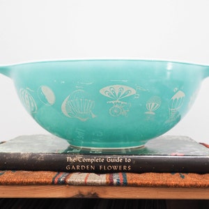 May include: A turquoise blue mixing bowl with a white design of hot air balloons. The bowl is sitting on top of a black book titled "The Complete Guide to Garden Flowers".