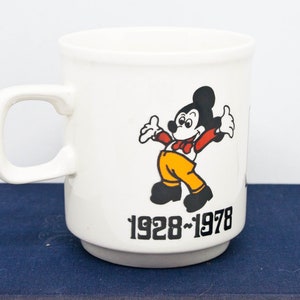 May include: White ceramic mug featuring a black and white image of Mickey Mouse wearing a red bow tie and yellow trousers. The mug has the text "1928-1978" printed on the bottom.