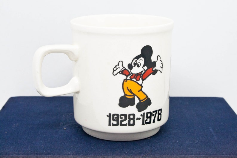 May include: White ceramic mug featuring a black and white image of Mickey Mouse wearing a red bow tie and yellow trousers. The mug has the text "1928-1978" printed on the bottom.
