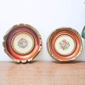 May include: Two circular wall plaques featuring a central cream-colored inlay with a colorful floral bouquet print. Each plaque is framed by a wide, deep orange-red border with a textured, scalloped gold-toned edge and a rope-like inner trim.