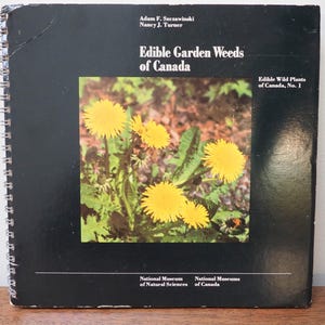 May include: A spiral-bound book titled "Edible Garden Weeds of Canada" with a black cover. The cover features a color photograph of yellow dandelions. The book is part of the "Edible Wild Plants of Canada" series.