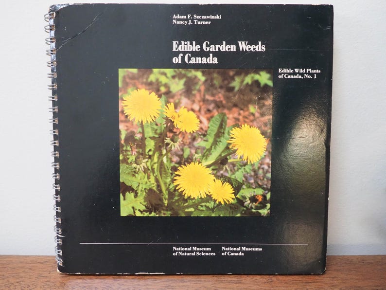 May include: A spiral-bound book titled "Edible Garden Weeds of Canada" with a black cover. The cover features a color photograph of yellow dandelions. The book is part of the "Edible Wild Plants of Canada" series.