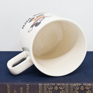 May include: White ceramic mug featuring a black and red cartoon character holding a baseball bat. The text "Ball Park" is printed on the mug.