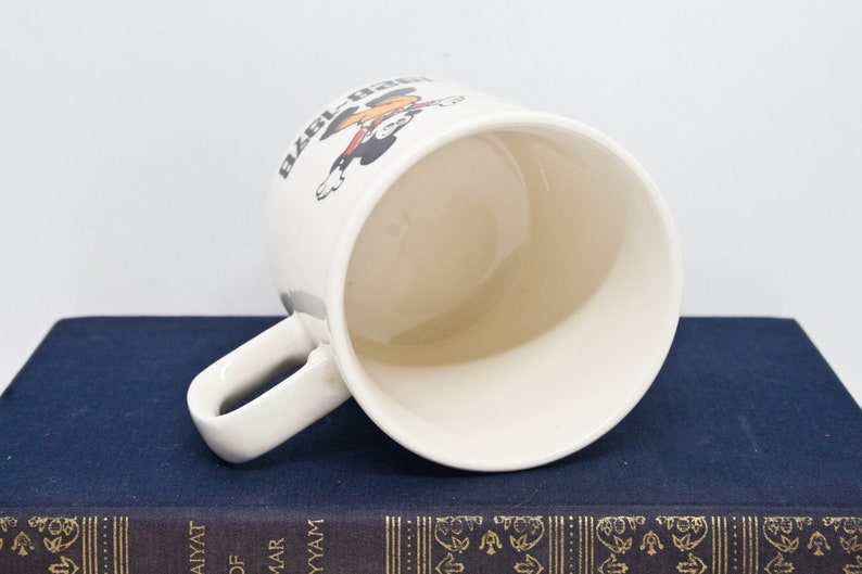 May include: White ceramic mug featuring a black and red cartoon character holding a baseball bat. The text "Ball Park" is printed on the mug.
