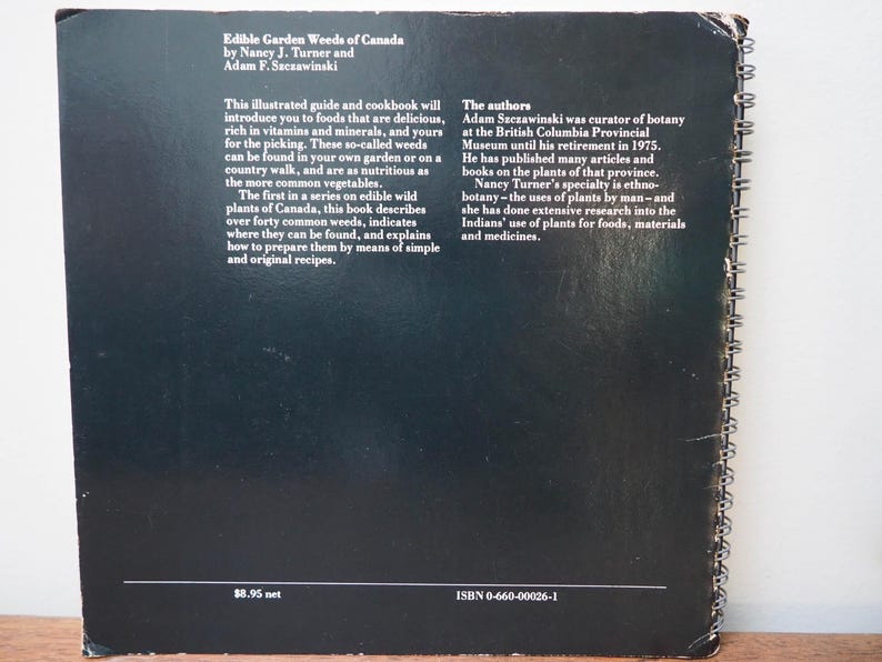 May include: A black book titled "Edible Garden Weeds of Canada" with white text. The book's text describes edible wild plants and recipes. The book has a spiral binding and is slightly worn.