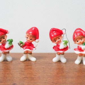 Op de afbeelding: Vier rode en witte kerst-elfen ornamenten met geschilderde gezichten en verschillende accessoires. De ornamenten zijn gemaakt van plastic en hebben een draadlus om op te hangen.