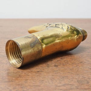 Puede incluir: Un tap&oacute;n de botella de lat&oacute;n vintage con forma de mano. La mano est&aacute; detallada con dise&ntilde;os grabados y tiene una base roscada para asegurarla a una botella. El lat&oacute;n tiene una p&aacute;tina c&aacute;lida y envejecida.