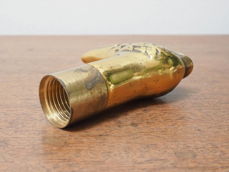 Puede incluir: Un tap&oacute;n de botella de lat&oacute;n vintage con forma de mano. La mano est&aacute; detallada con dise&ntilde;os grabados y tiene una base roscada para asegurarla a una botella. El lat&oacute;n tiene una p&aacute;tina c&aacute;lida y envejecida.