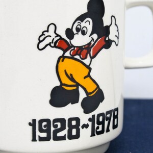 May include: White ceramic mug featuring a black and white illustration of Mickey Mouse wearing a red bow tie and yellow trousers. The mug has the text "1928-1978" printed on the front.