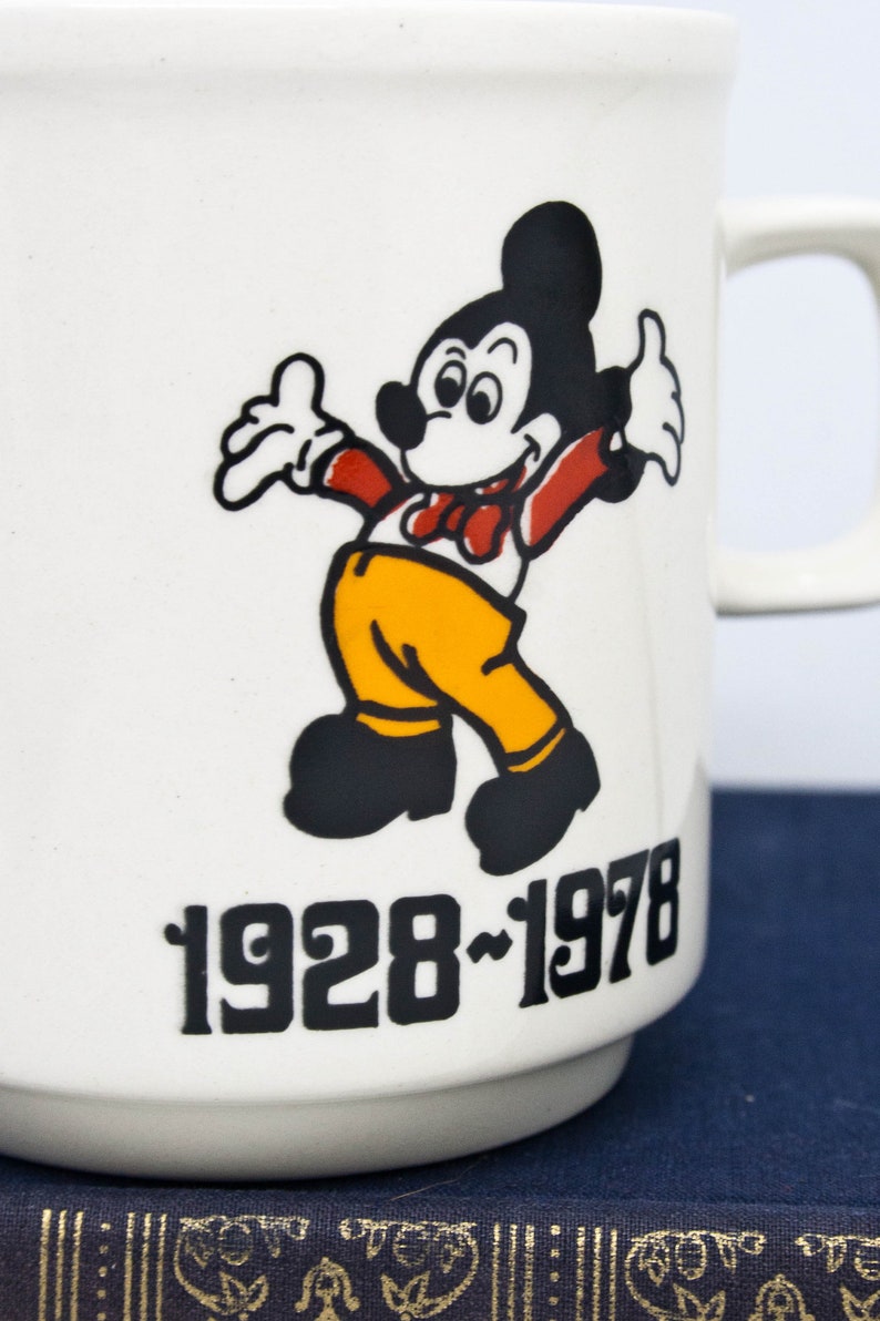 May include: White ceramic mug featuring a black and white illustration of Mickey Mouse wearing a red bow tie and yellow trousers. The mug has the text "1928-1978" printed on the front.