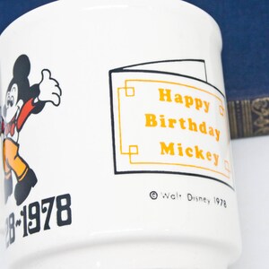May include: White ceramic mug featuring a black and white illustration of Mickey Mouse. The mug has orange text that reads "Happy Birthday Mickey" and "&copy; Walt Disney 1978". The bottom of the mug has black text that says "28-1978".