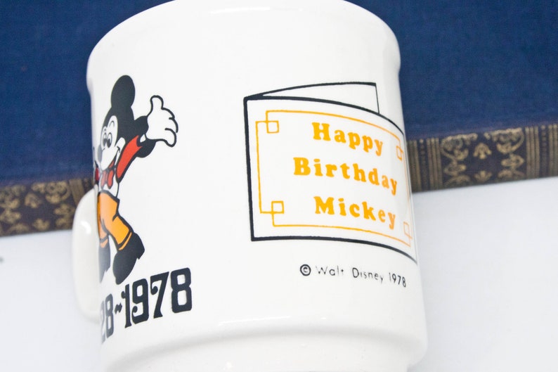 May include: White ceramic mug featuring a black and white illustration of Mickey Mouse. The mug has orange text that reads "Happy Birthday Mickey" and "&copy; Walt Disney 1978". The bottom of the mug has black text that says "28-1978".