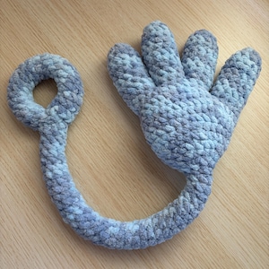 May include: A soft, plush, light blue and gray crocheted toy shaped like a hand with a loop. The hand has four fingers and a thumb. The loop is connected to the hand by a long, curved piece. The toy is made of a textured yarn.