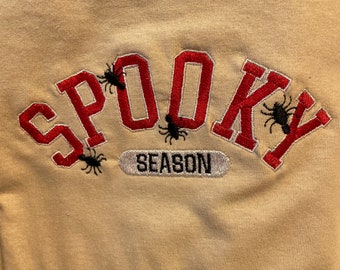 May include: A beige sweatshirt with red embroidered text that reads "Spooky Season" with black spider silhouettes.
