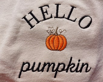 May include: A beige sweatshirt with black embroidered text that reads "Hello pumpkin". The text is arched over a small orange embroidered pumpkin.