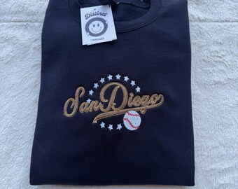 May include: A navy blue sweatshirt with the embroidered text "San Diego" in a script font, surrounded by stars and a baseball. A tag with a smiley face logo is attached.