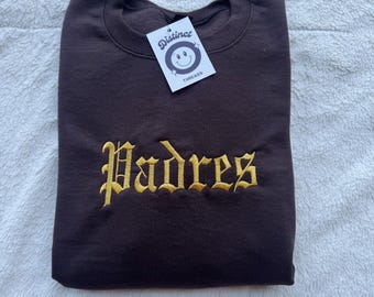 May include: A dark brown sweatshirt with the word "Padres" embroidered in gold lettering. A white tag with the "Distinct Threads" logo is attached to the sweatshirt.