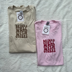 May include: Two t-shirts with the text "Drinks Well With Others" in a retro font. The shirt on the left is beige with brown text. The shirt on the right is pink with burgundy text.