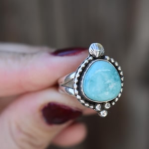 Könnte beinhalten: Ein silberner Ring mit einem großen, hellblauen Edelstein. Der Ring hat einen detaillierten, perlenbesetzten Rand und einen kleinen muschelförmigen Akzent. Der Edelstein hat einen Durchmesser von etwa 2 cm. Der Ring wird vor einem unscharfen Hintergrund gehalten.