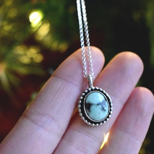 Puede incluir: Un collar de plata con un colgante ovalado. El colgante tiene una piedra verde claro y marrón, enmarcada por un borde plateado con cuentas. El collar se muestra sobre un fondo verde borroso.