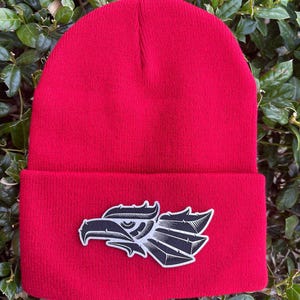 May include: Red beanie with a black and white eagle graphic on the front. The eagle is a stylized design with a sharp beak and feathers.