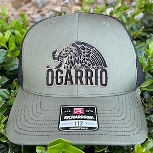 May include: A green and black trucker hat with a black eagle graphic and the text "OGARRIO" in a tan color. The hat is adjustable and has a black mesh back. The hat has a Richardson label with the text "EST. 1970 ORIGINAL 112 TRUCKER ADJUSTABLE".