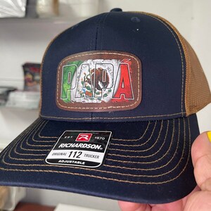 May include: A navy blue and brown trucker hat with a brown leather patch featuring the word "PAPA" in green, white, and red, with the Mexican flag in the background. The hat has a brown mesh back and a black adjustable strap with the text "EST. 1970 RICHARDSON 112 ORIGINAL TRUCKER ADJUSTABLE".