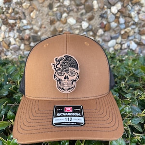 May include: A brown and black trucker hat with a black and white skull and eagle design. The hat has an adjustable strap and a label that reads "Richardson 112 Original Trucker Adjustable".