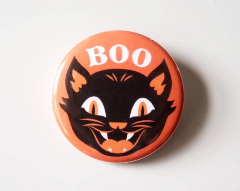 May include: A round orange button with a black cat face and the word "BOO" in white. The cat has orange ears, eyes, and mouth, with white fangs. A Halloween-themed design.