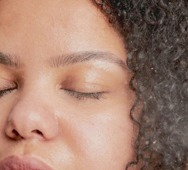 May include: Close-up of a person's face with their eyes closed. The person has brown skin and dark curly hair. The skin is smooth and appears to be glowing.