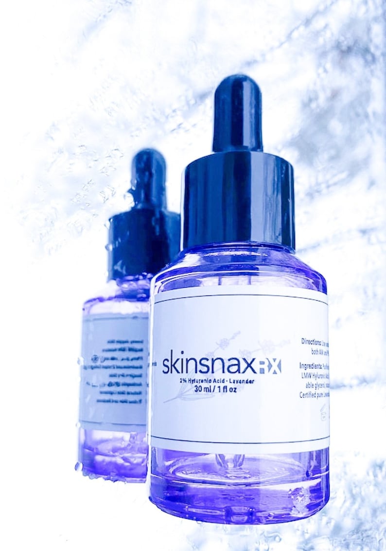 May include: Two clear glass bottles with blue dropper tops filled with a purple liquid. The bottles are labelled "skinsnax 2% Hyaluronic Acid - Lavender 30 ml / 1 fl oz".