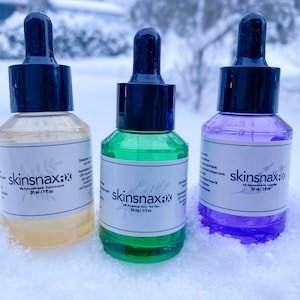 May include: Three glass dropper bottles with labels that say "skinsnax" and list different ingredients. The bottles are sitting on a snowy surface.