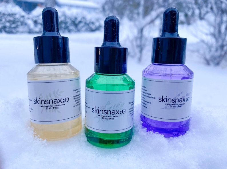 May include: Three glass dropper bottles with labels that say "skinsnax" and list different ingredients. The bottles are sitting on a snowy surface.