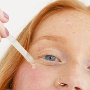 May include: A close-up of a person's face with a clear dropper bottle being applied to the skin under the eye. The person has a nose ring and freckles.