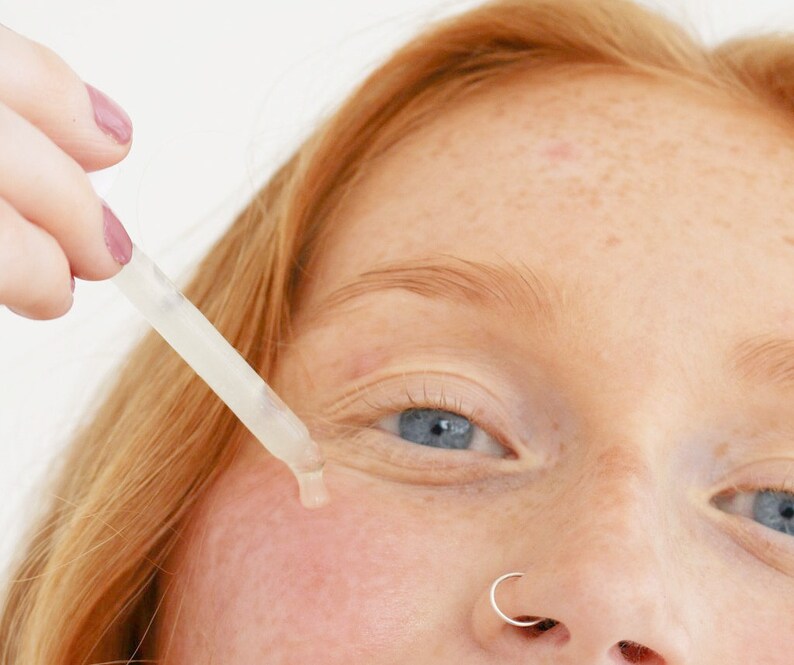 May include: A close-up of a person's face with a clear dropper bottle being applied to the skin under the eye. The person has a nose ring and freckles.
