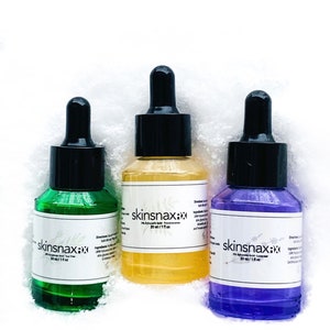 May include: Three glass bottles with dropper tops containing skin care serums. The bottles are labelled "skinsnax:xx" and contain 2% Hyaluronic Acid. The serums are green, yellow, and purple.
