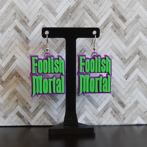 May include: A pair of earrings with the words "Foolish Mortal" in green lettering on a purple background. The earrings are hanging from silver hooks and are displayed on a black stand.