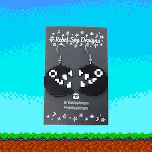 Może przedstawiać: Para czarnych, pikselowych kolczyków Pac-Mana z białymi detalami. Kolczyki wiszą na srebrnych haczykach. Kolczyki znajdują się na czarnej kartce z tekstem "Rebel Spy Designs" i uchwytami mediów społecznościowych.