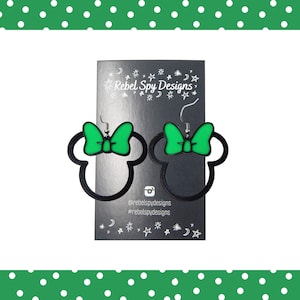 May include: A pair of black and green Minnie Mouse-shaped earrings. The earrings have a black outline of the mouse head with a green bow. They are displayed on a black card with the text "Rebel Spy Designs".