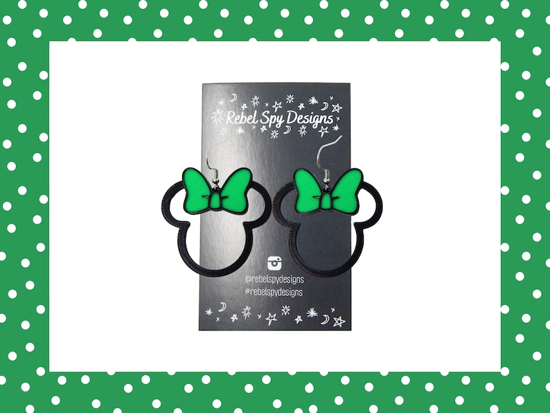 May include: A pair of black and green Minnie Mouse-shaped earrings. The earrings have a black outline of the mouse head with a green bow. They are displayed on a black card with the text "Rebel Spy Designs".