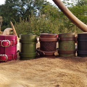 May include: Seven leather-wrapped cups with different designs and colours. The cups are lined up in a row on a furry surface.