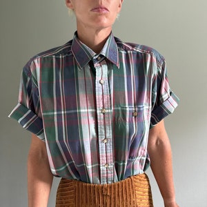 May include: A short-sleeved button-down shirt with a plaid pattern in shades of green, blue, pink, and white. The shirt has a classic collar and a button-up front.