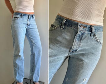 May include: Light blue denim jeans with distressed detailing. The jeans feature a classic five-pocket design and a straight leg fit. The jeans are worn with a white vest top and black and white heels.