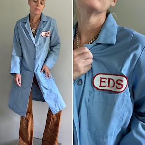 May include: A light blue work coat featuring a pointed collar, long sleeves, and a button-down front. The garment includes two large lower pockets and one chest pocket with an oval patch displaying the red text EDS on a white background.