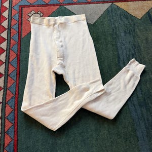 May include: Cream-coloured thermal underwear bottoms with a waffle knit texture. The long johns have a button-up fly and ribbed cuffs at the ankles. The garment is laid out on a green and red patterned rug.