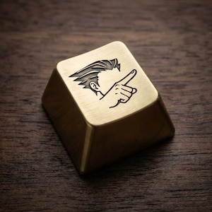 May include: A brass-coloured, square keycap featuring a stylized illustration of a person pointing. The design includes a detailed outline of a hairstyle, ear, and hand. The keycap has a trapezoidal shape.