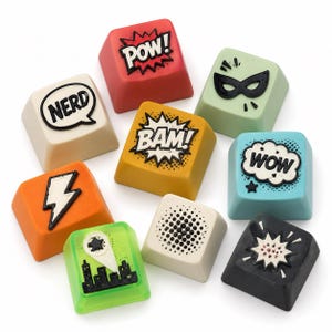 May include: A collection of colorful keyboard keycaps with comic book-style graphics. Keycaps include the words "POW!", "BAM!", "WOW!", and "NERD", along with other designs like a lightning bolt and a cityscape.