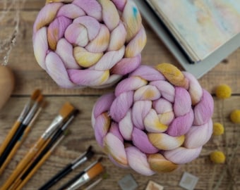 May include: Two thick, braided coils of soft wool roving featuring a variegated color palette of pastel pink, lavender, and golden yellow. The fibers are densely packed and twisted into circular shapes, showcasing a smooth, fluffy texture.