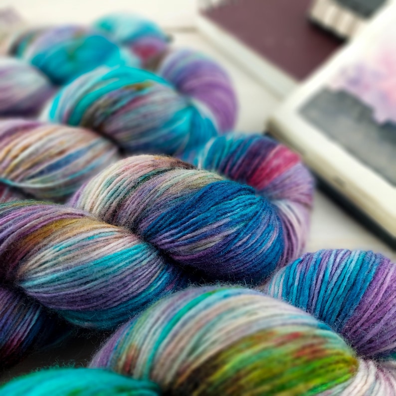 May include: Close-up of several skeins of colorful yarn in shades of blue, purple, and teal. The yarn is tightly wound and appears soft. A notebook is visible in the background. The yarn is likely used for knitting or crocheting projects.